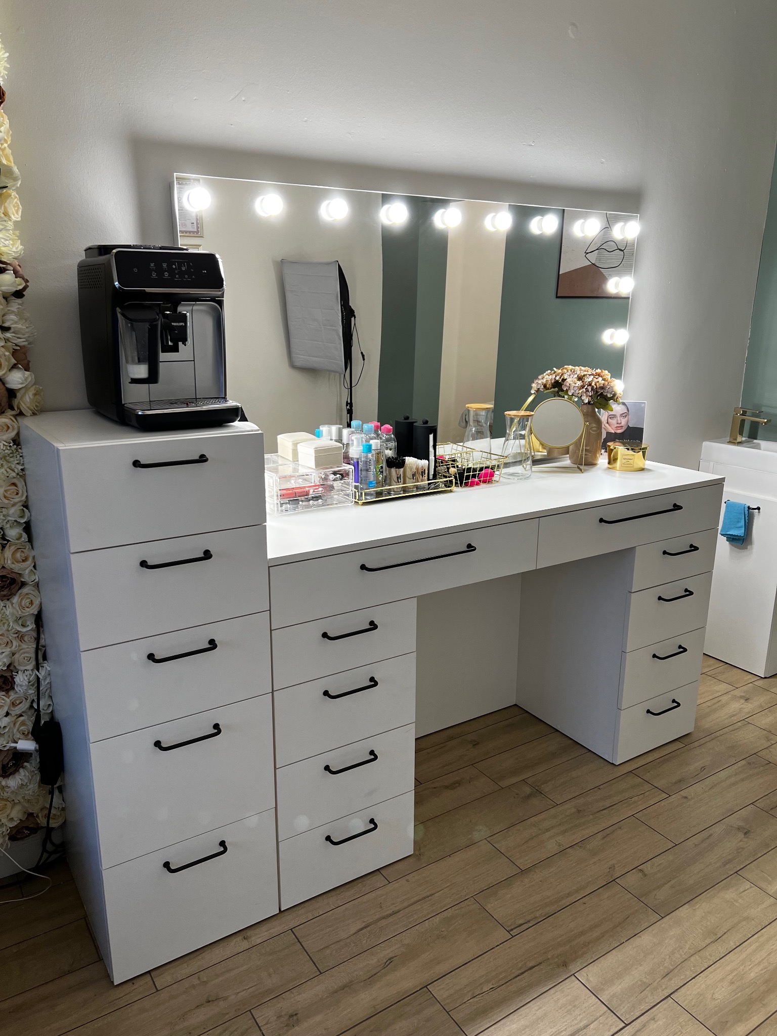 Biała toaletka z szufladami i lustrem otoczonym żarówkami, na blacie akcesoria do makijażu, ekspres do kawy, dekoracja kwiatowa i drewniana podłoga.