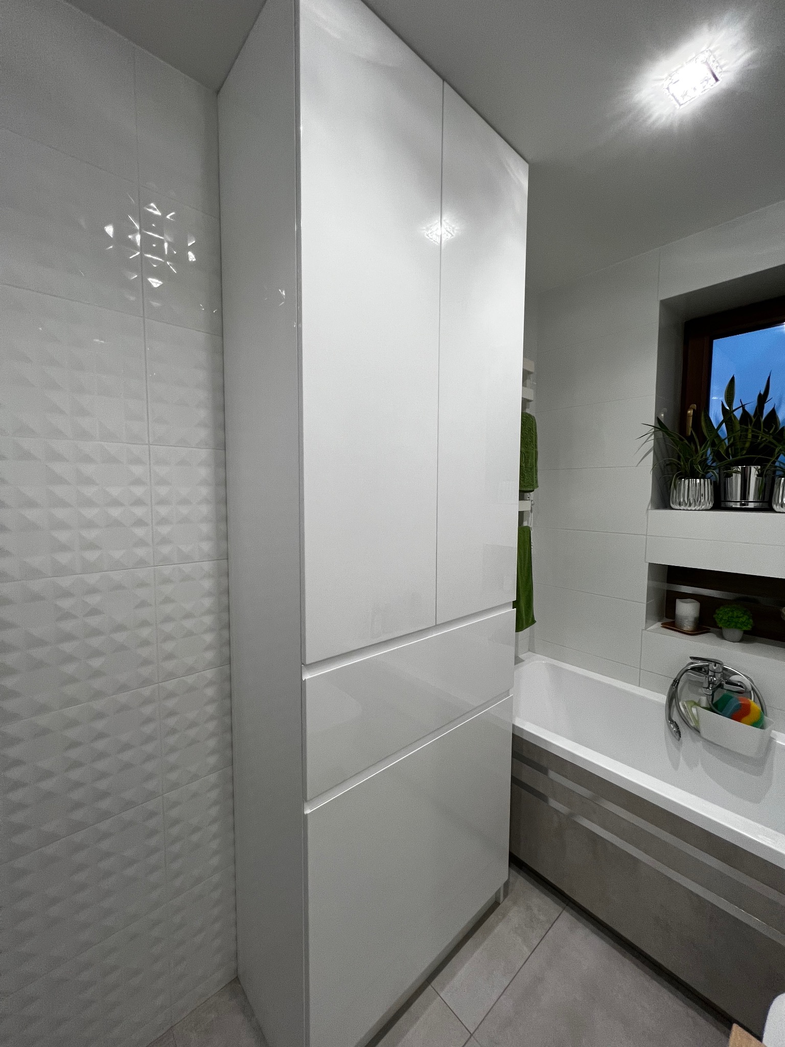 Wysoka, biała szafa łazienkowa z frontami bez uchwytów, umieszczona obok wanny w nowoczesnej łazience z białymi płytkami o trójkątnym wzorze i oknem z widokiem na zewnątrz.