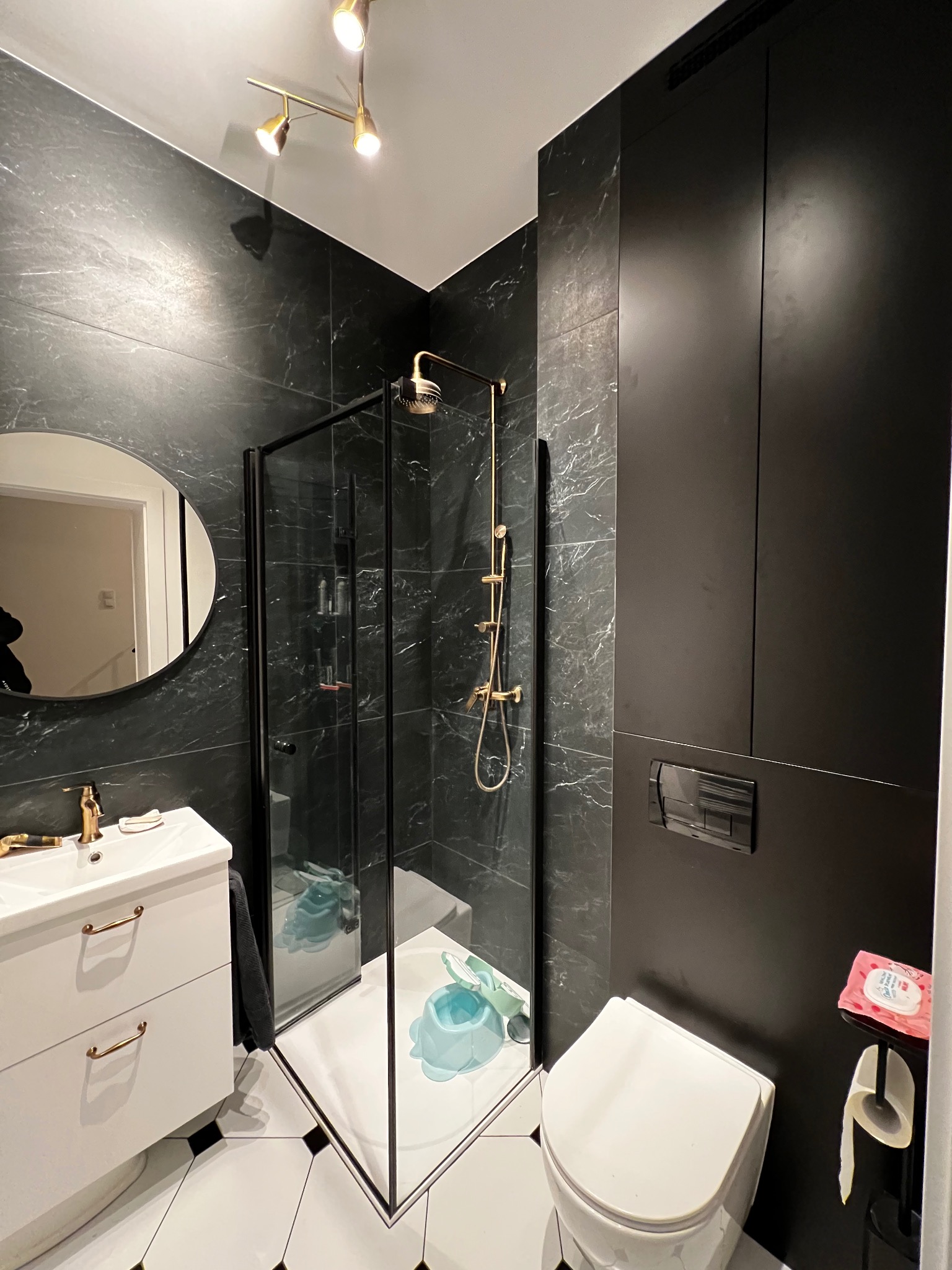 Nowoczesna łazienka z czarnymi marmurowymi płytkami, kabiną prysznicową z czarną ramą i złotą armaturą, białą szafką pod umywalkę z złotymi uchwytami i okrągłym lustrem.