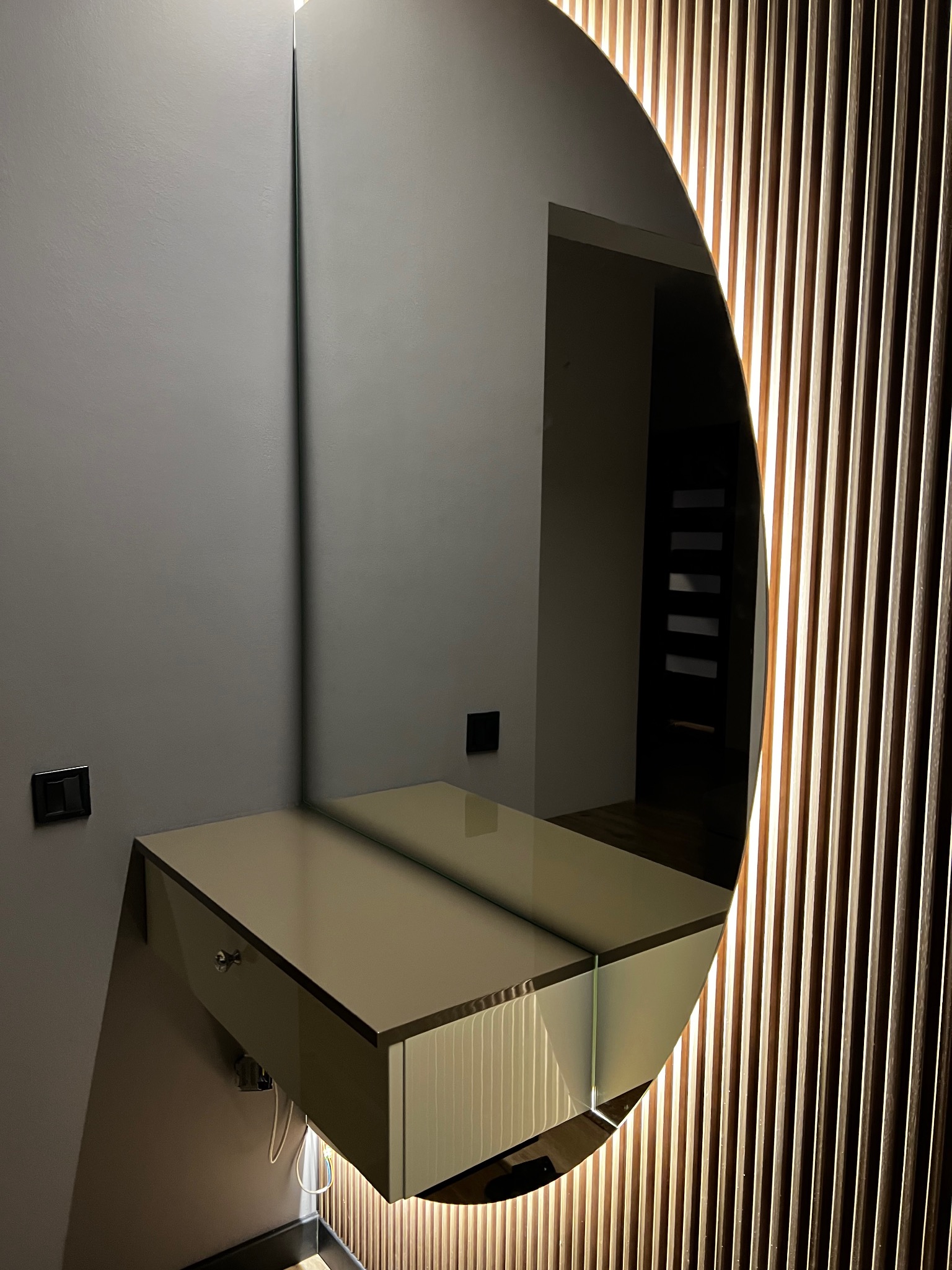 Nowoczesna toaletka z szufladą i okrągłym lustrem z podświetleniem LED, zamontowana na szarej ścianie, obok drewnianej ściany z pionowych listew.