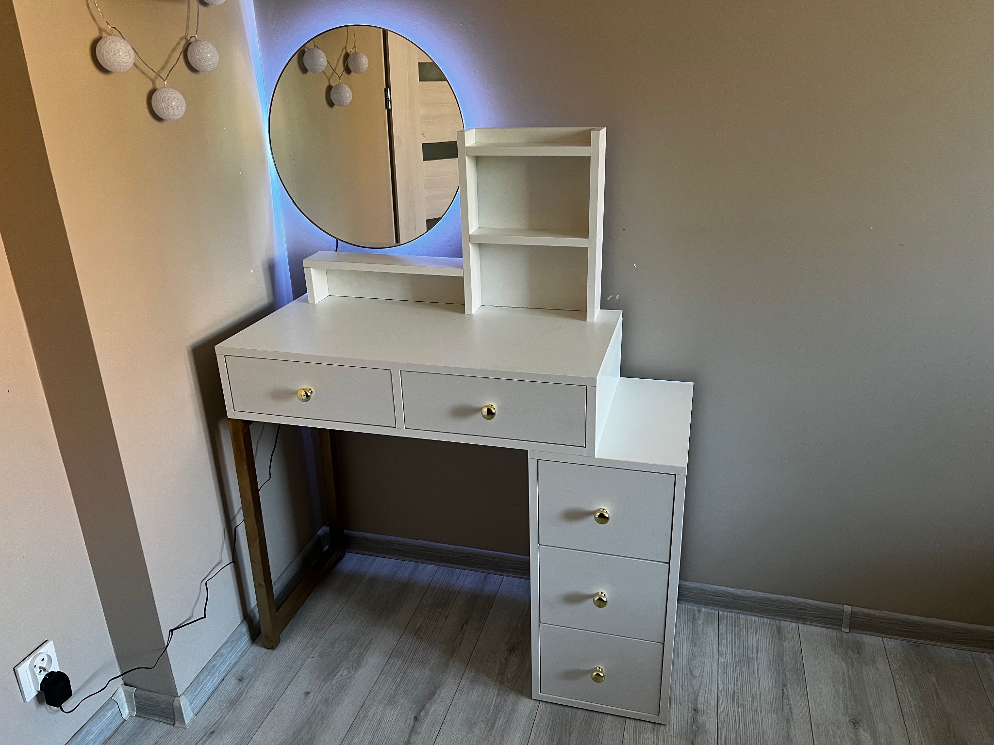 Biała toaletka z trzema szufladami po prawej stronie i trzema szufladami pod blatem, okrągłe lustro z niebieskim podświetleniem LED, złote gałki szuflad, ustawiona na szarym panelu podłogowym.