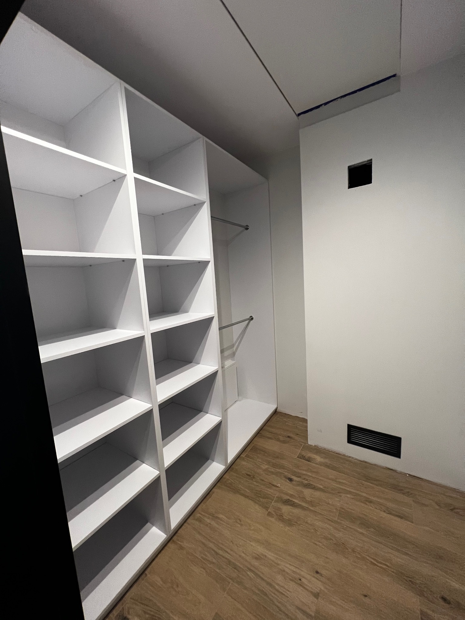 Wnętrze garderoby z białą zabudową szafy na wymiar, widoczne półki i drążek na ubrania, podłoga wykończona panelami imitującymi drewno.