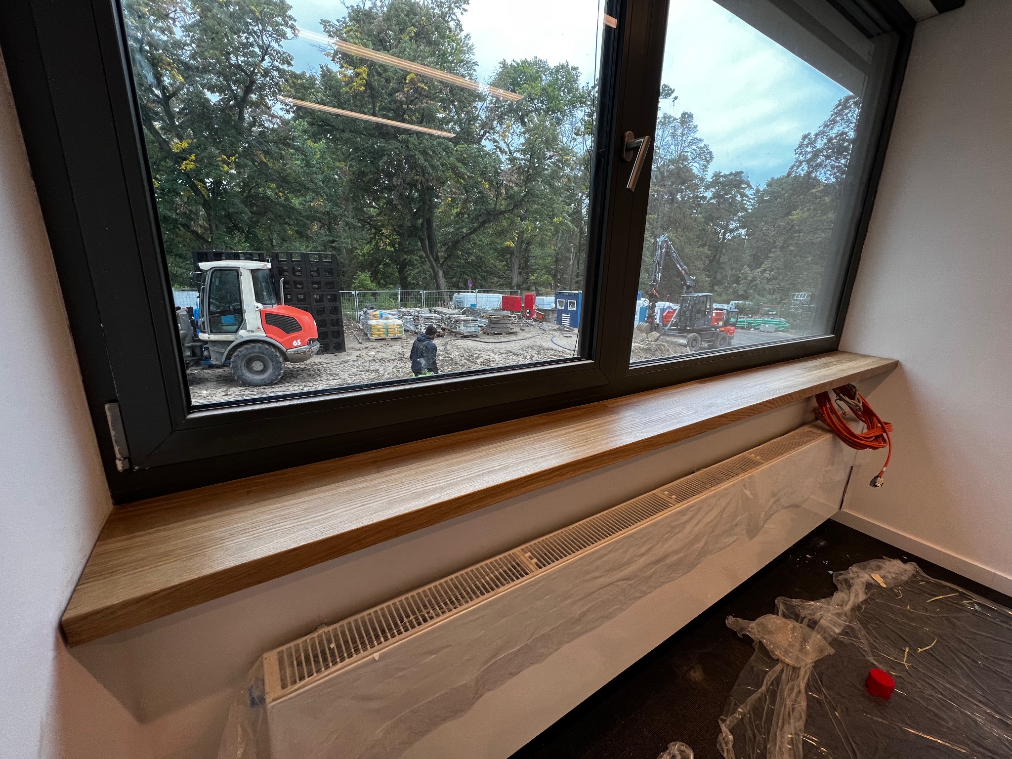 Wnętrze pokoju z drewnianym parapetem i grzejnikiem, widok na plac budowy za oknem.