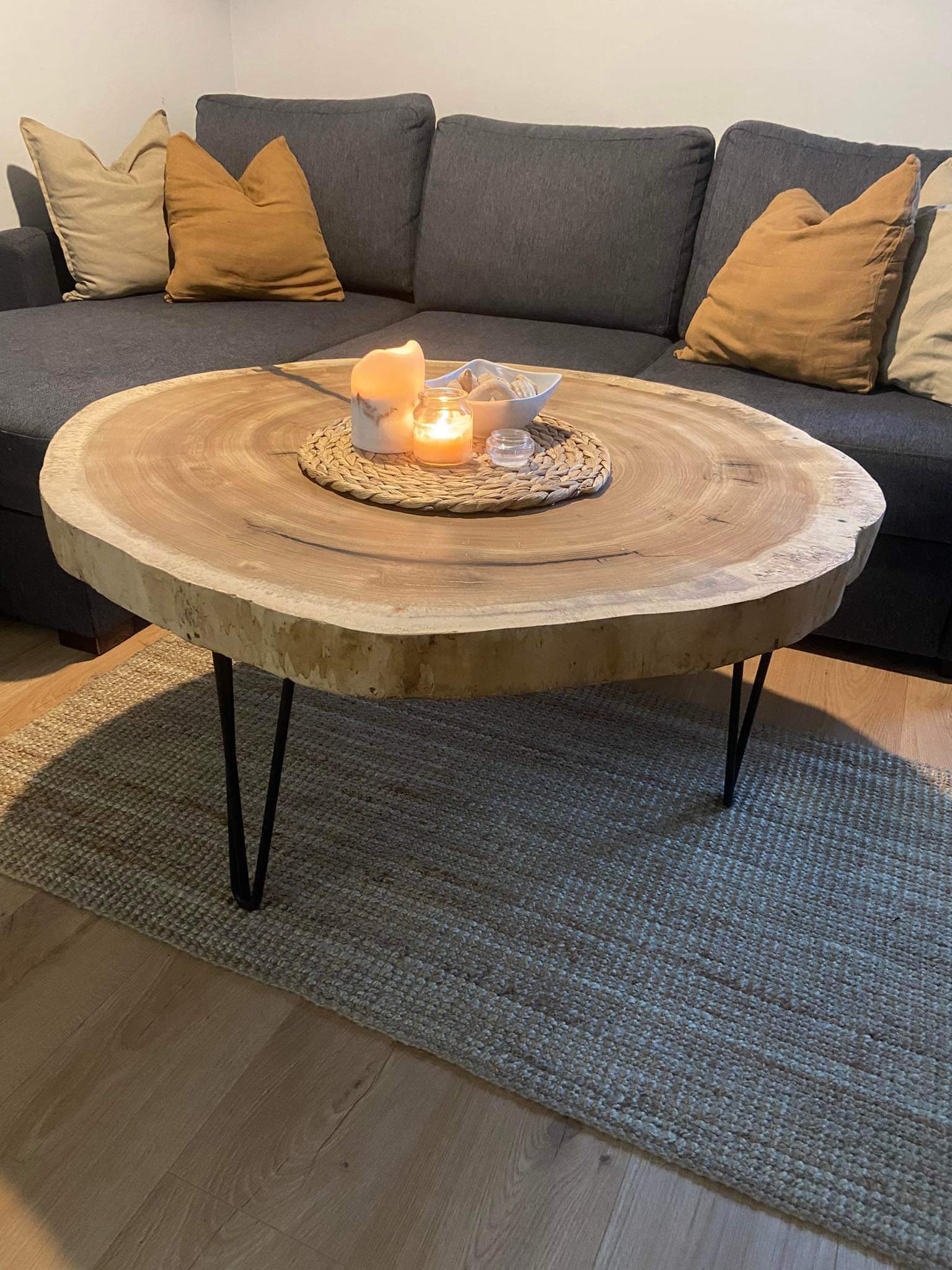 Stolik kawowy z blatem z litego drewna o nieregularnym kształcie, ustawiony na metalowych nogach, z dekoracjami w postaci świec i muszelek, na tle szarej sofy i jutowego dywanu.