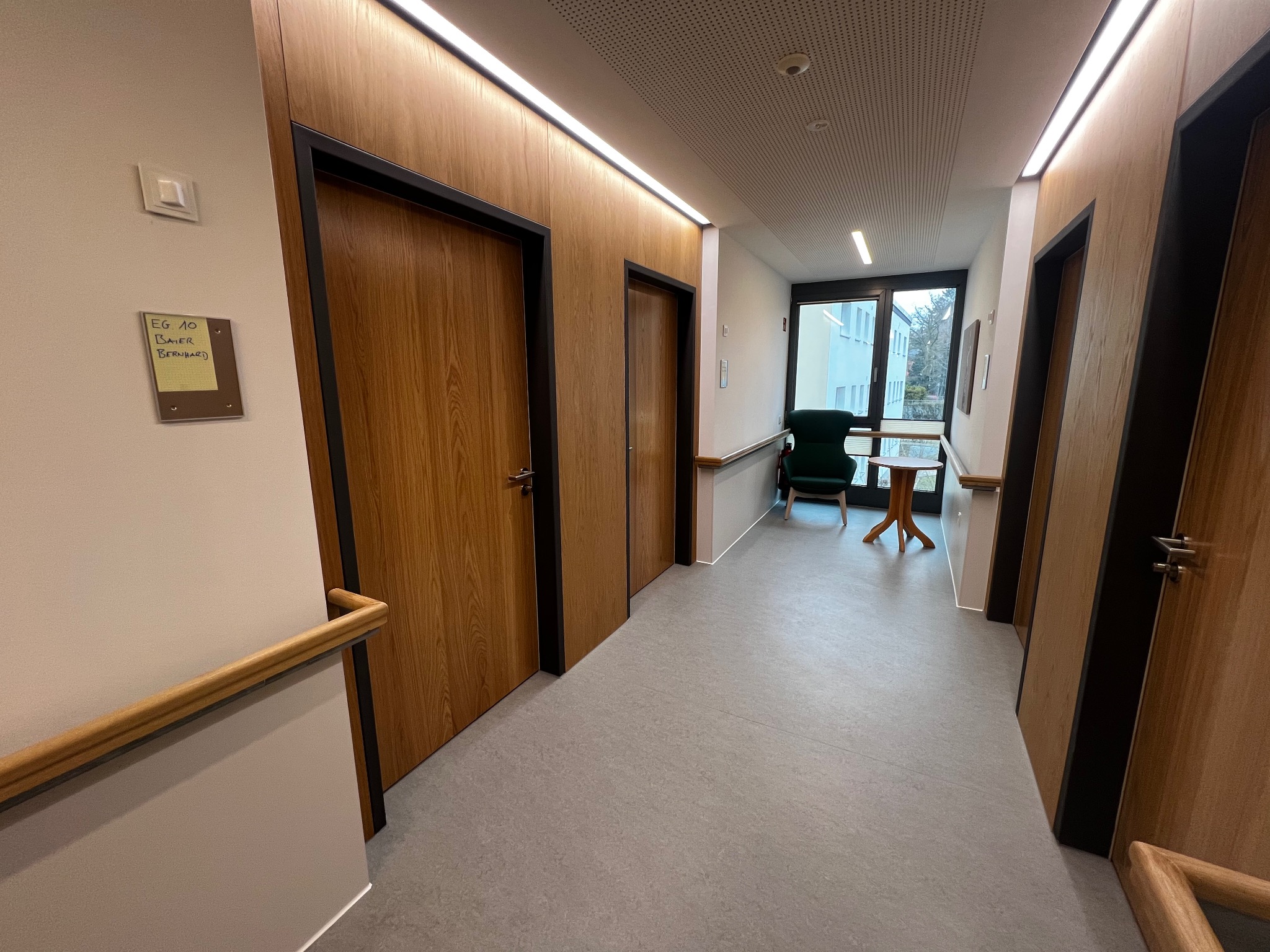 Korytarz z drewnianymi drzwiami z czarnymi ościeżnicami, drewnianą poręczą i widokiem na zewnątrz przez okno na końcu korytarza.