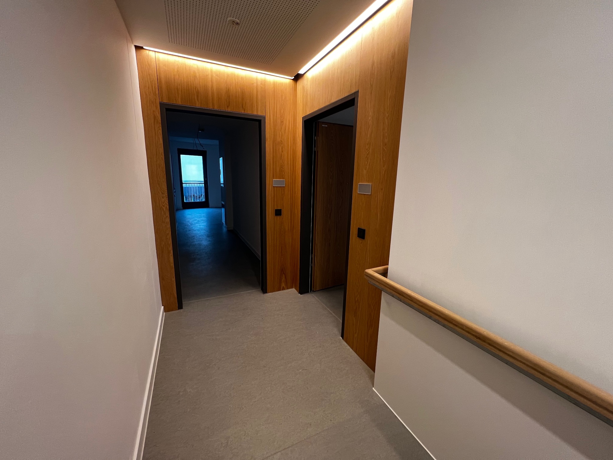 Korytarz z dwoma drewnianymi drzwiami z czarnymi ościeżnicami, podświetlony listwą LED, z widoczną balustradą schodową i fragmentem balkonu w oddali.