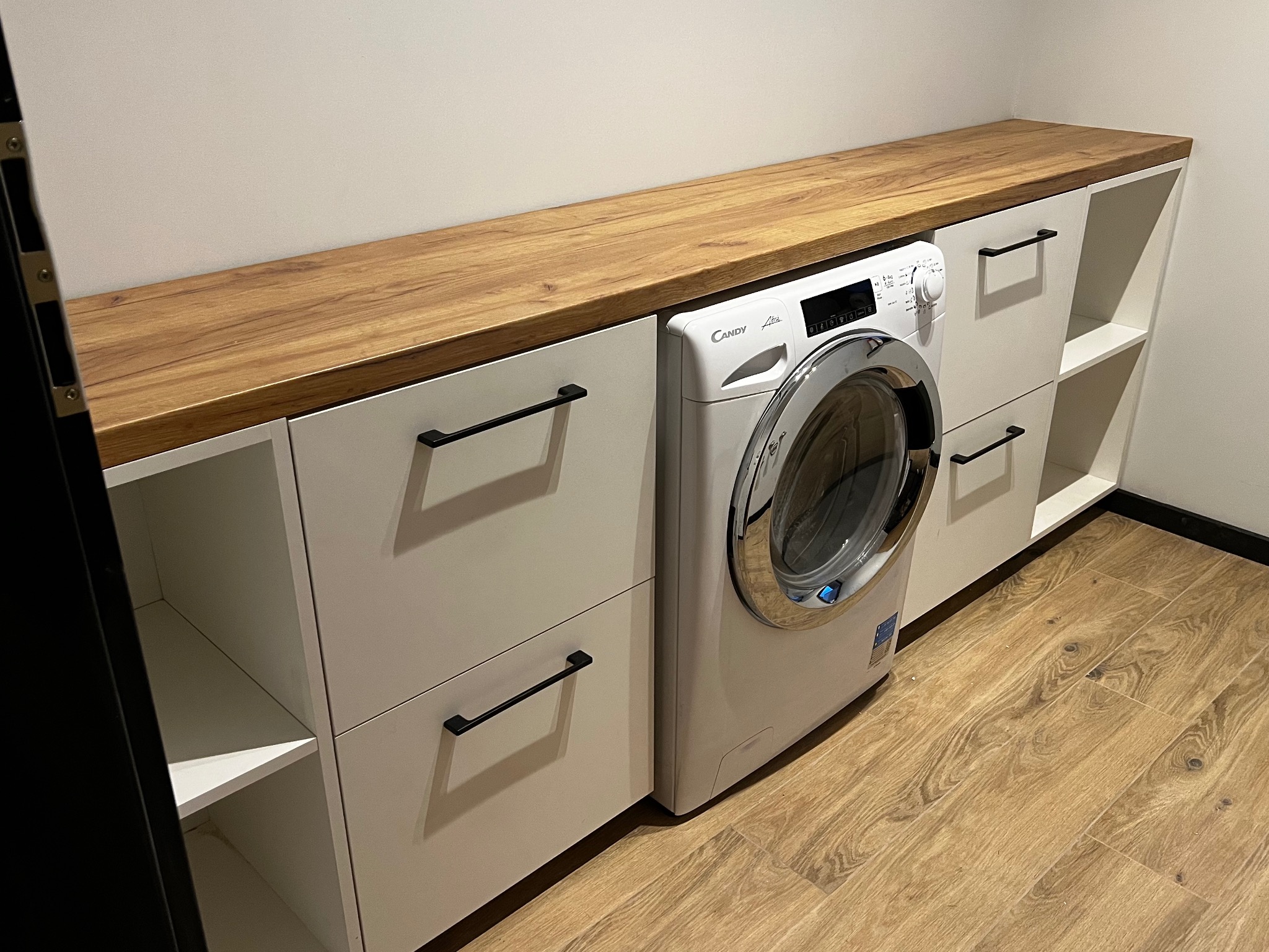 Białe szafki z drewnianym blatem, wkomponowane w przestrzeń z pralką Candy, czarne uchwyty, podłoga imitująca drewno.