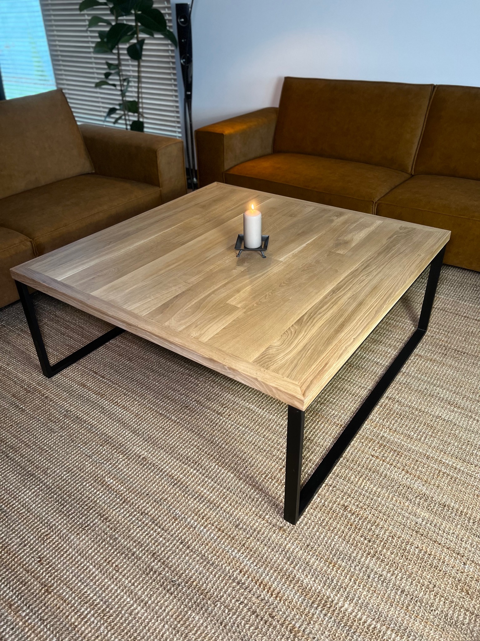 Kwadratowy stolik kawowy z jasnego drewna z czarnymi, metalowymi nogami, na tle brązowej sofy i dywanu ze sznurka, z zapaloną świecą na blacie.