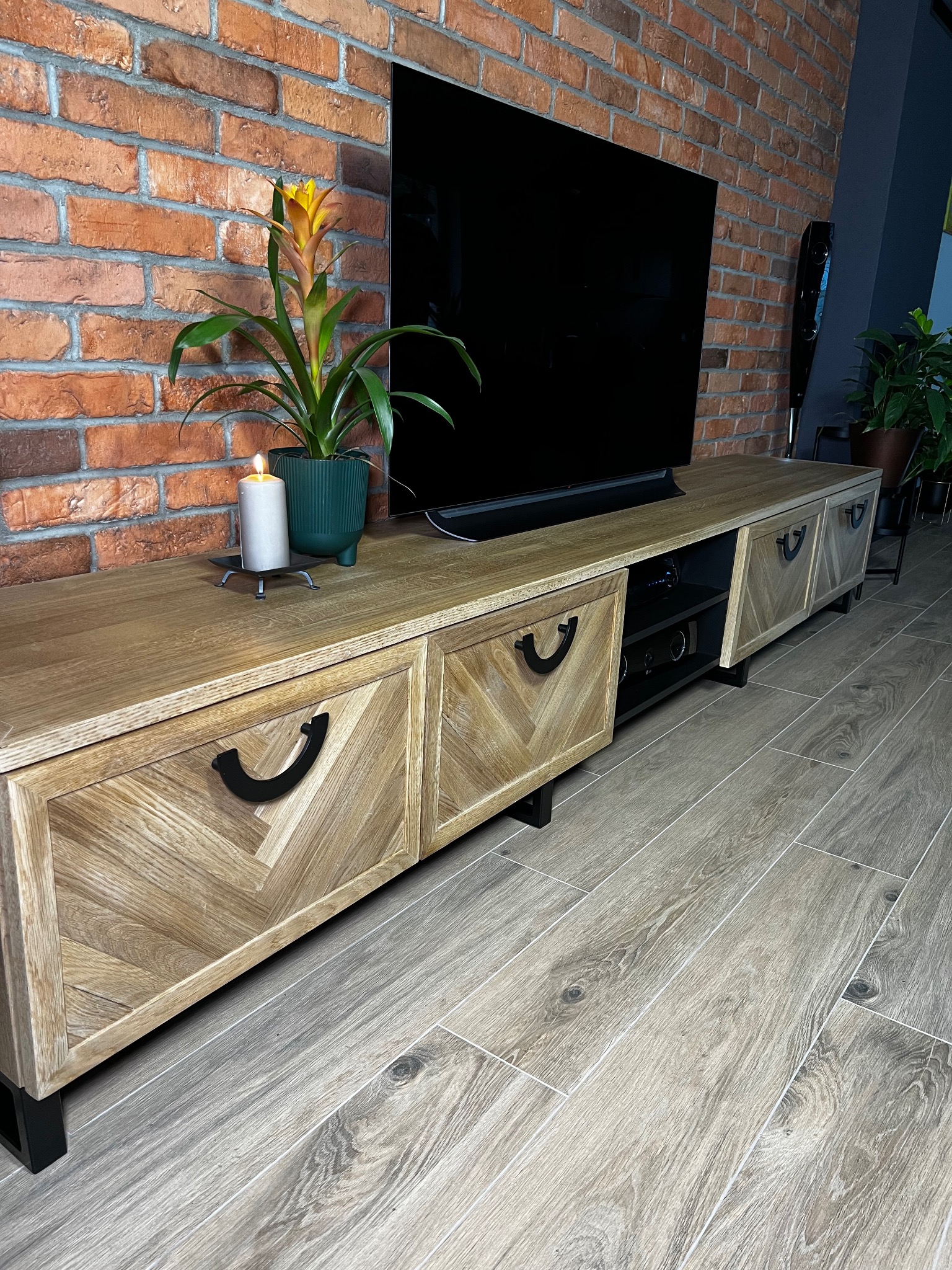 Drewniana szafka RTV z motywem jodełki na frontach, czarne uchwyty, stojąca na czarnych nóżkach, na blacie świeca i roślina w doniczce, na tle ściany z czerwonej cegły i telewizora.