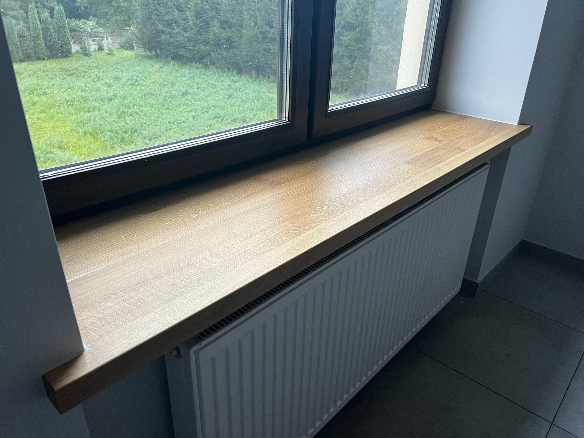 Jasny, drewniany parapet wewnętrzny z widokiem na zielony trawnik przez okno z ciemną ramą, pod parapetem grzejnik.