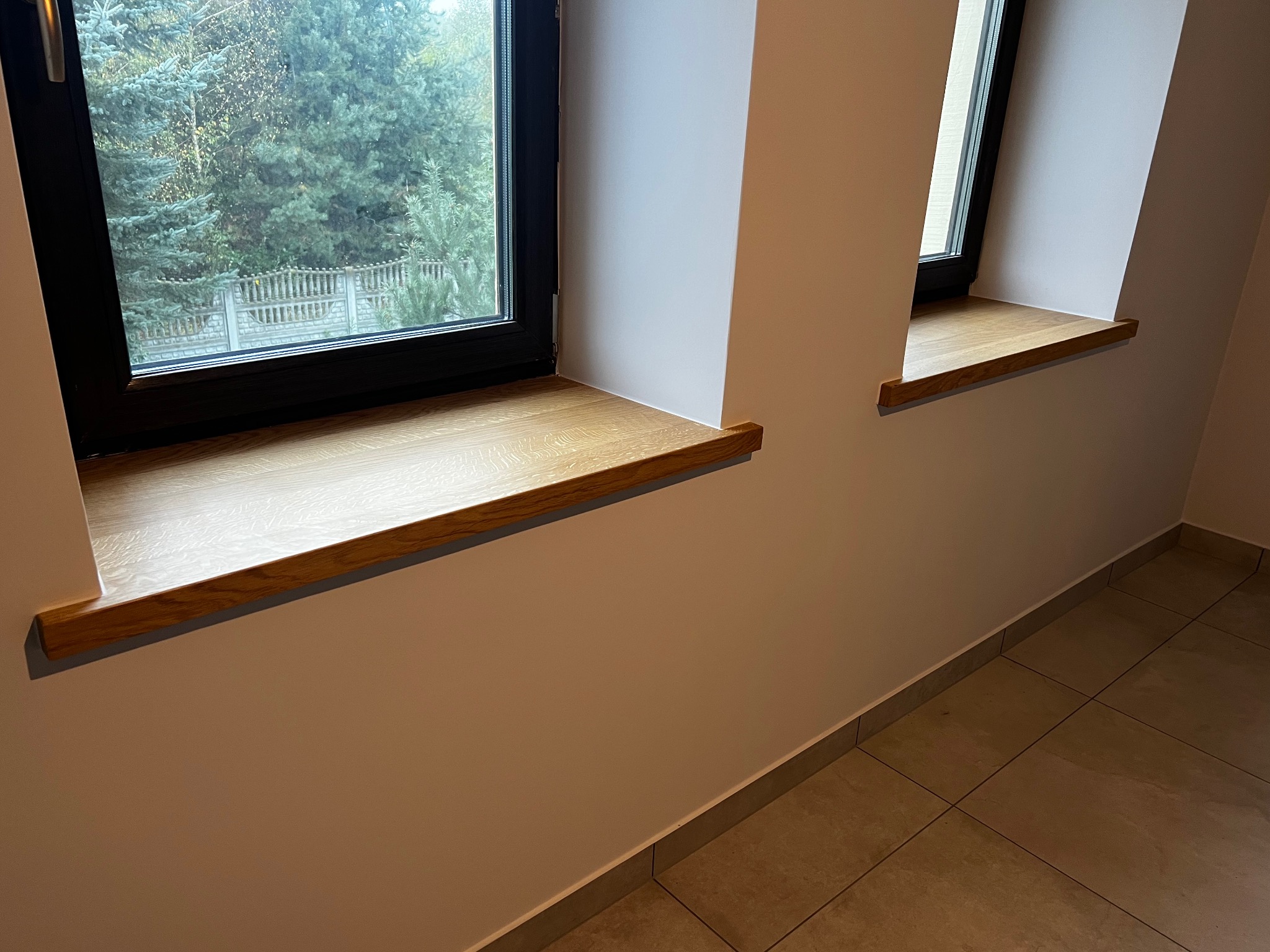Dwa drewniane parapety okienne o jasnym odcieniu drewna, zamontowane w oknach z czarnymi ramami na tle jasnych ścian i płytek podłogowych.