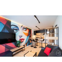 F.U.H. SZYMON REMONTUJE - Wnętrze mieszkania z dużym, geometrycznym muralem przedstawiającym twarz kobiety, drewniana podłoga, minimalistyczne meble, czarne oświetlenie punktowe i aneks kuchenny z wysokimi stołkami barowymi.