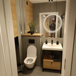 F.U.H. SZYMON REMONTUJE - Nowoczesna łazienka w odcieniach szarości i drewna, z wiszącą toaletą, umywalką na metalowym stelażu z drewnianą szafką i okrągłym lustrem z podświetleniem LED, w którym odbija się osoba robiąca...