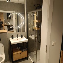 F.U.H. SZYMON REMONTUJE - Nowoczesna łazienka w szarościach z kabiną prysznicową, umywalką na szafce z drewnianym frontem i okrągłą lampą LED, widoczna toaleta i fragment kuchni w odbiciu lustra.