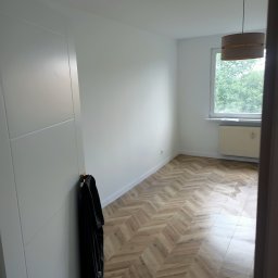 F.U.H. SZYMON REMONTUJE - Pusty pokój po remoncie z parkietem ułożonym w jodełkę, świeżo pomalowanymi białymi ścianami, grzejnikiem pod oknem i wiszącą lampą z abażurem, z czarną kurtką wiszącą na drzwiach.