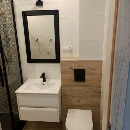 F.U.H. SZYMON REMONTUJE - Nowoczesna łazienka z białymi płytkami, drewnopodobnymi panelami, wiszącą toaletą i czarną armaturą, widok na umywalkę z szafką i lustro z oświetleniem.