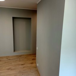 F.U.H. SZYMON REMONTUJE - Wnętrze pokoju po remoncie: szare ściany, drewnopodobne panele podłogowe, wnęka w ścianie pomalowana na szaro, widoczne listwy przypodłogowe.