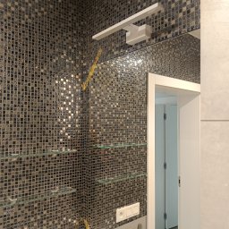 F.U.H. SZYMON REMONTUJE - Mozaika z drobnych, błyszczących płytek w odcieniach szarości i srebra, szklane półki, oświetlenie nad lustrem oraz widoczne gniazdka elektryczne w łazience, z fragmentem żółtej taśmy malarskiej.