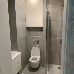 F.U.H. SZYMON REMONTUJE - Nowoczesna łazienka w odcieniach szarości z białymi meblami: wisząca szafka, umywalka z szafką i toaleta, kabina prysznicowa z metalową ramą.