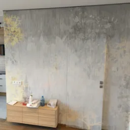 Ściana z ukrytymi drzwiami pokryta szarą tapetą z motywem roślinnym w odcieniach złota i bieli, z drewnianą szafką i akcesoriami malarskimi u dołu.