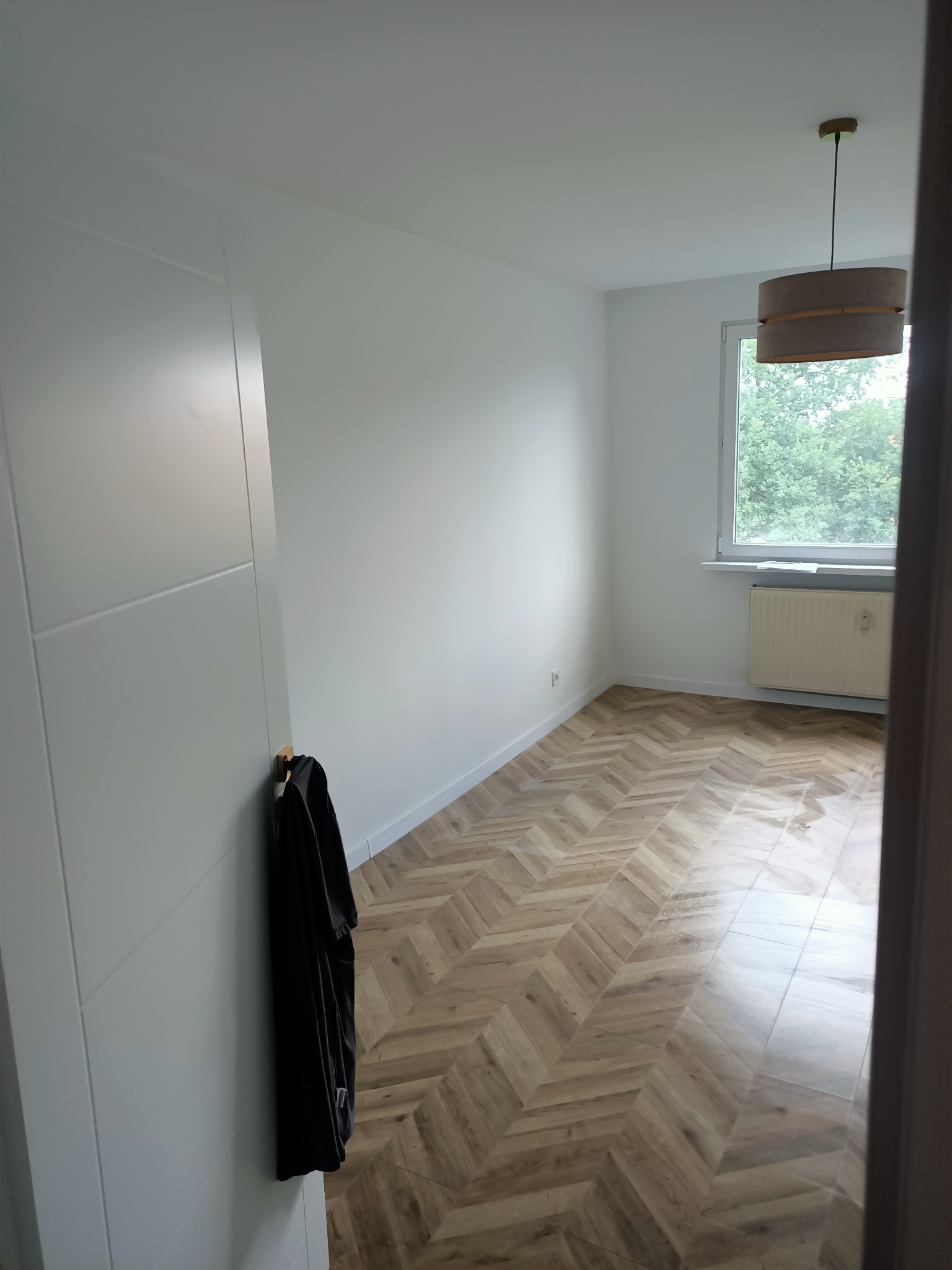 Pusty pokój po remoncie z parkietem ułożonym w jodełkę, świeżo pomalowanymi białymi ścianami, grzejnikiem pod oknem i wiszącą lampą z abażurem, z czarną kurtką wiszącą na drzwiach.