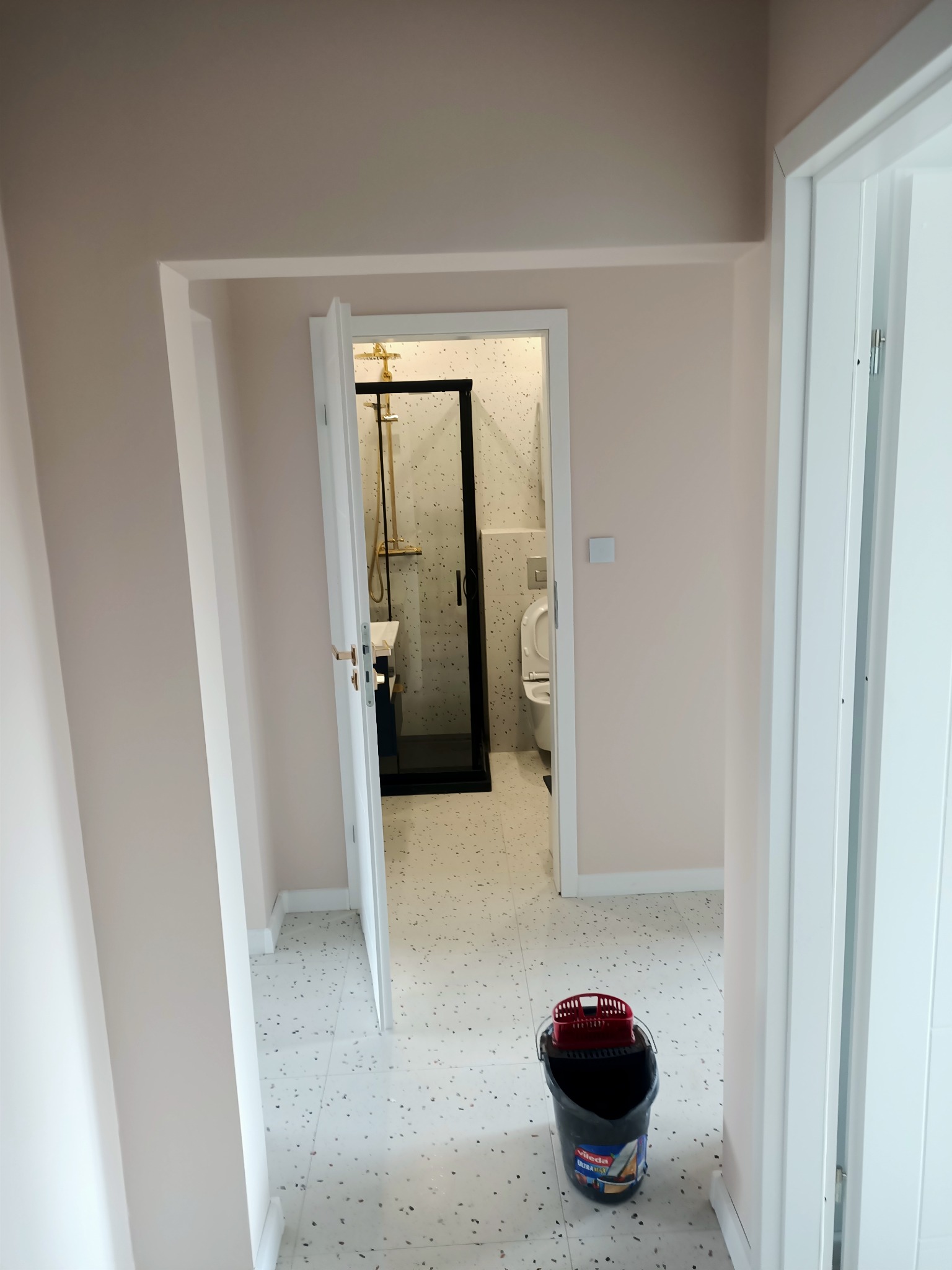 Widok na wykończoną łazienkę z kabiną prysznicową z czarną ramą i złotą armaturą, widoczny przez otwarte drzwi, na pierwszym planie wiadro z mopem.