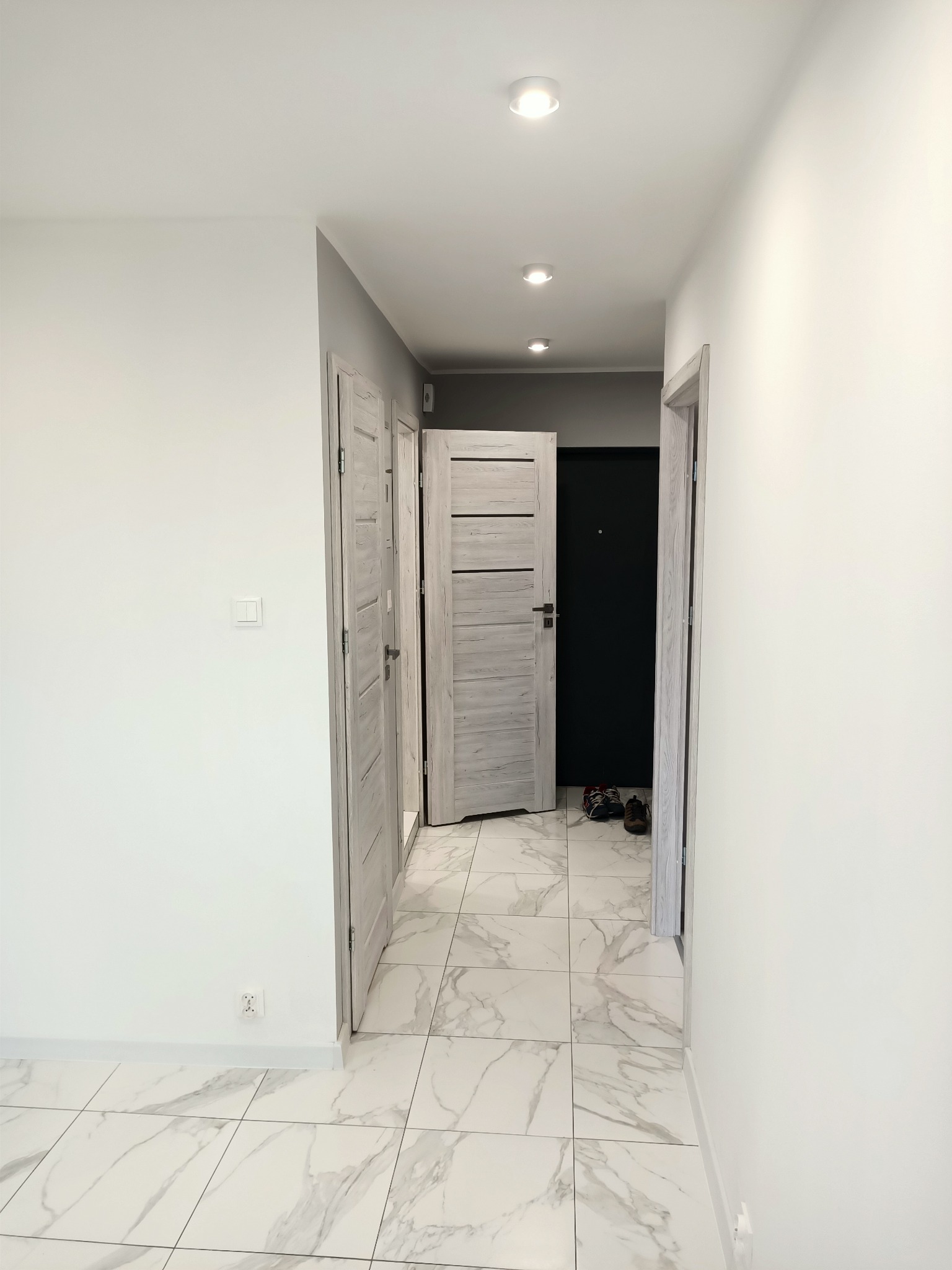 Korytarz z białymi ścianami, marmurową podłogą i drewnianymi drzwiami, widoczne oświetlenie punktowe na suficie.