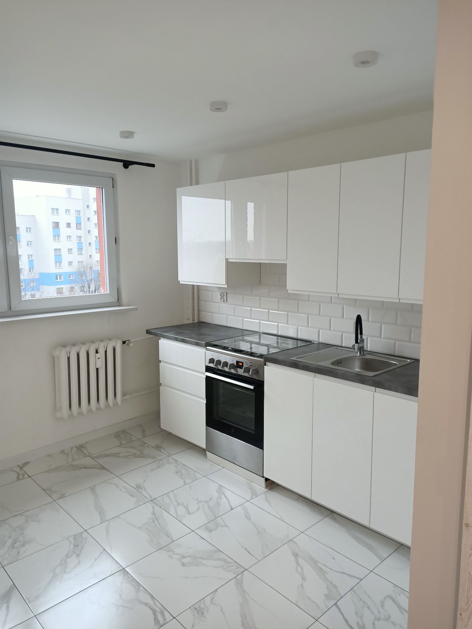 Jasna kuchnia z białymi, błyszczącymi szafkami, szarym blatem i marmurową podłogą, widok na blok z okna.