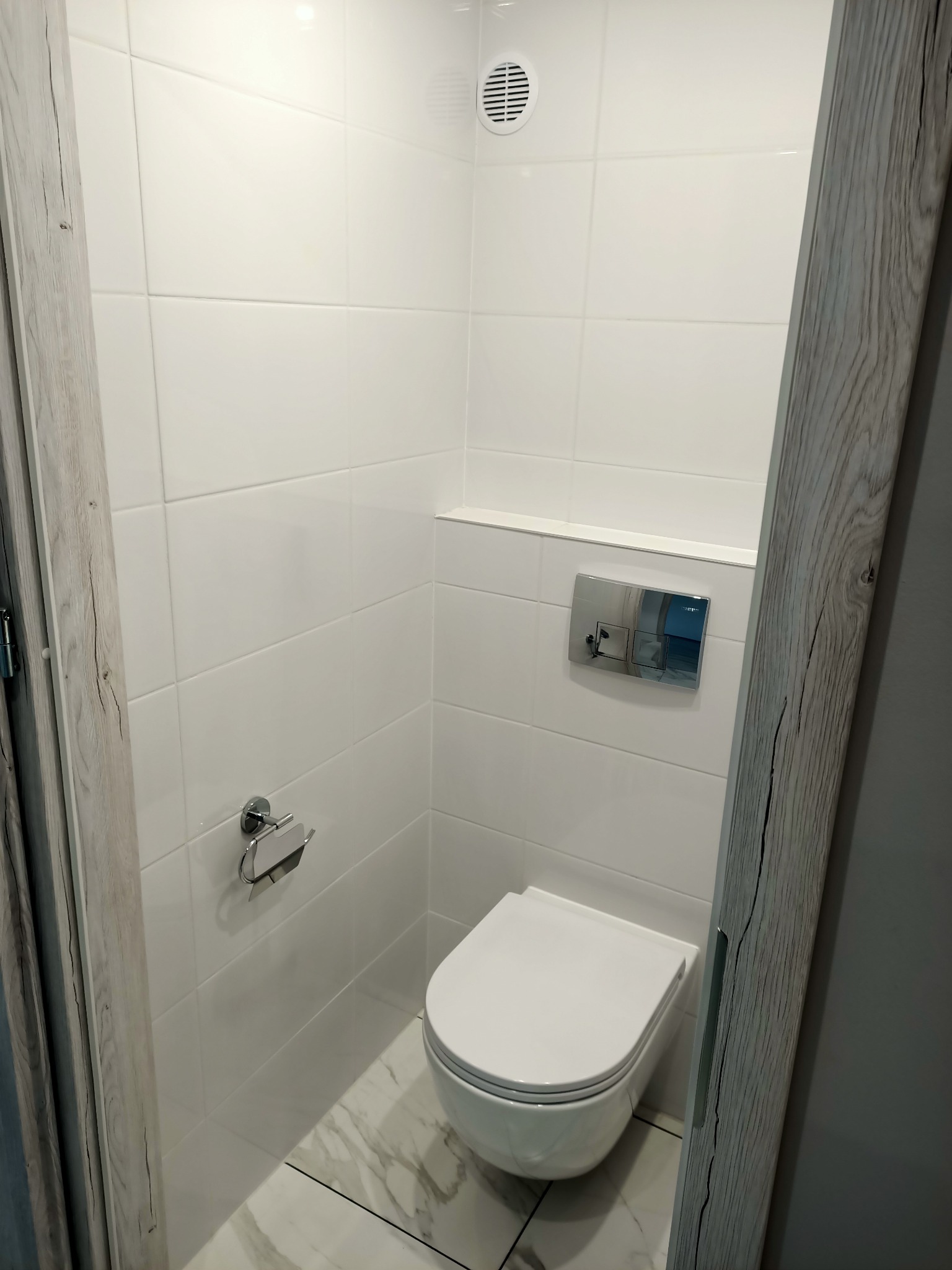 Nowoczesna toaleta w małej łazience, białe płytki na ścianach, podłoga imitująca marmur, rama drzwi z efektem surowego drewna.