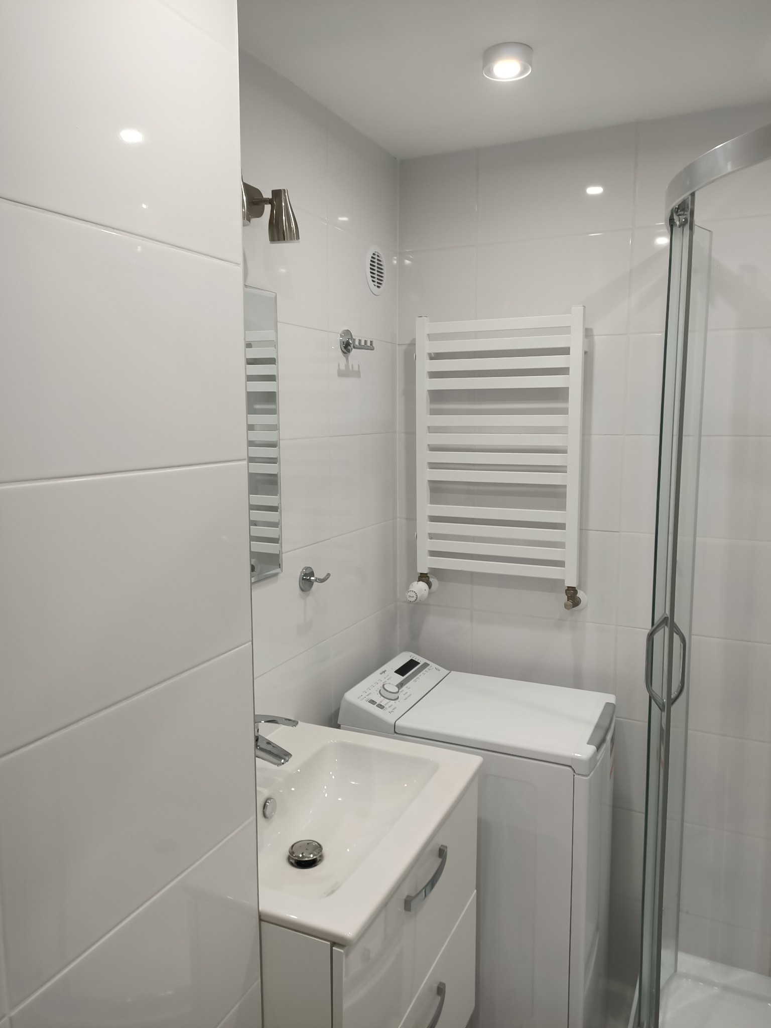 Biała łazienka z kabiną prysznicową, umywalką z szafką, pralką i grzejnikiem drabinkowym, wykończona białymi płytkami.
