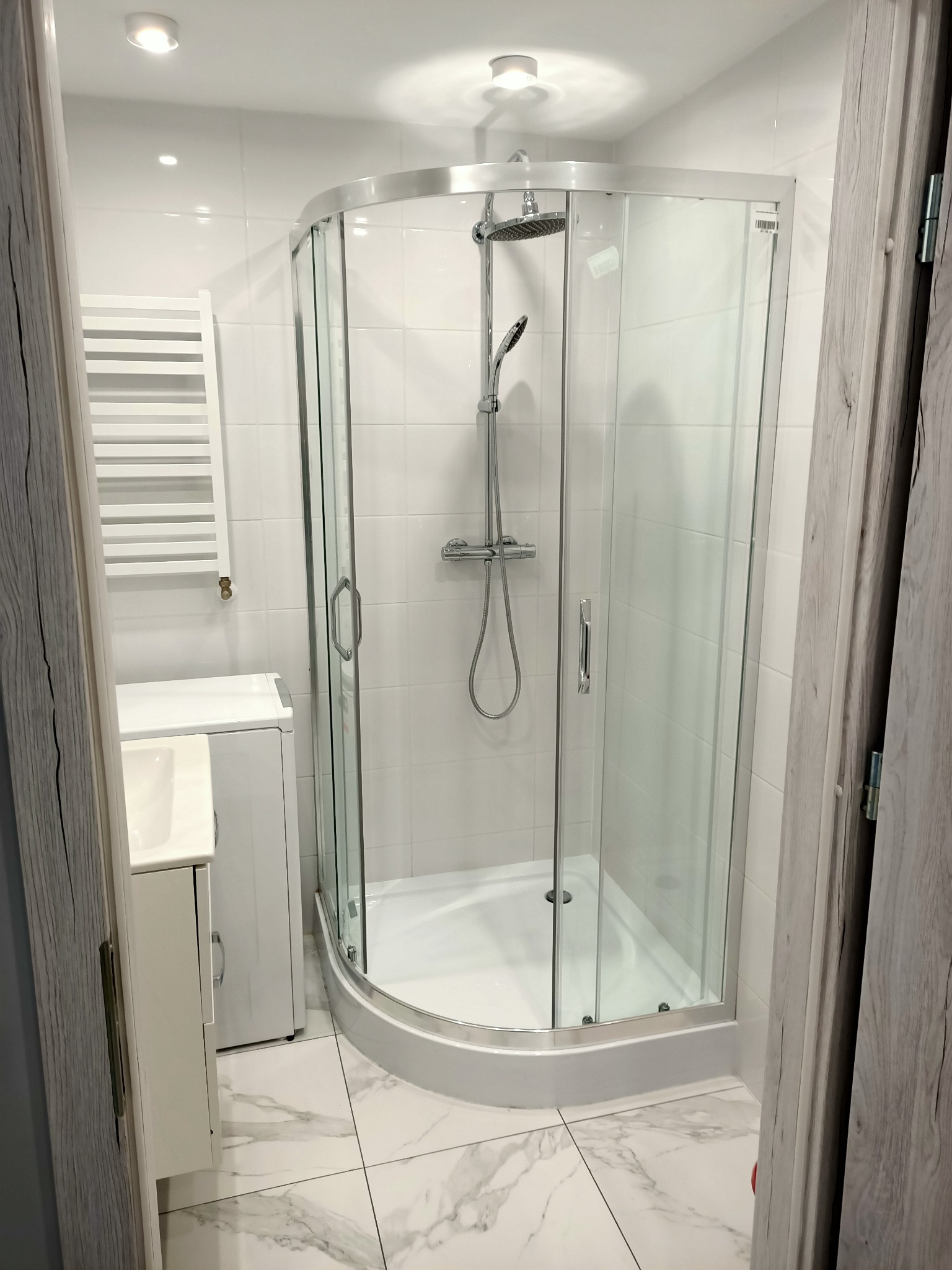 Nowoczesna kabina prysznicowa z chromowaną armaturą, białe płytki na ścianach i podłodze z marmurowym wzorem, widok przez drewnopodobne futryny drzwi.