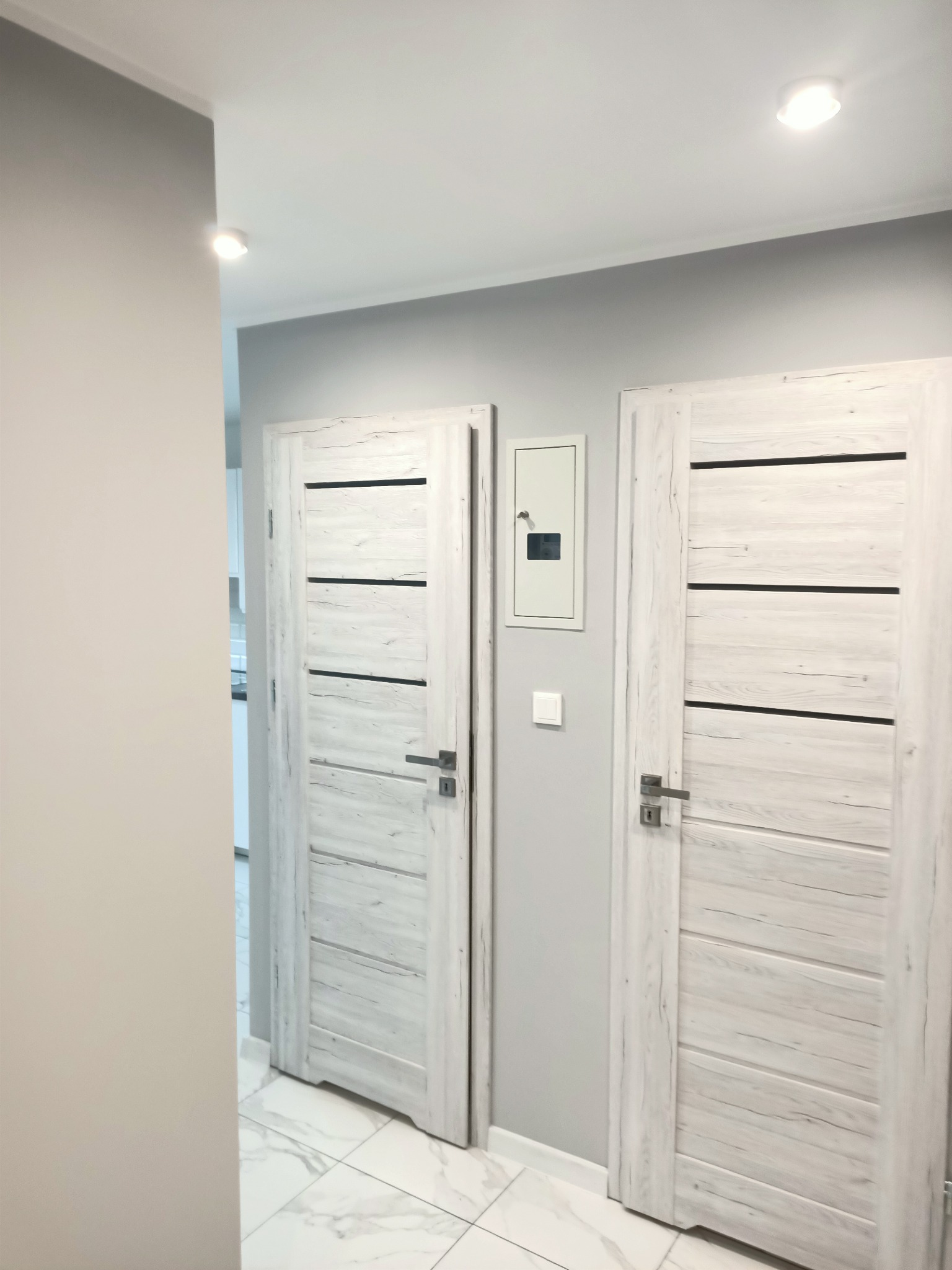 Nowoczesny przedpokój z dwoma białymi drzwiami o drewnianej fakturze, szarą ścianą z skrzynką elektryczną i marmurową podłogą.