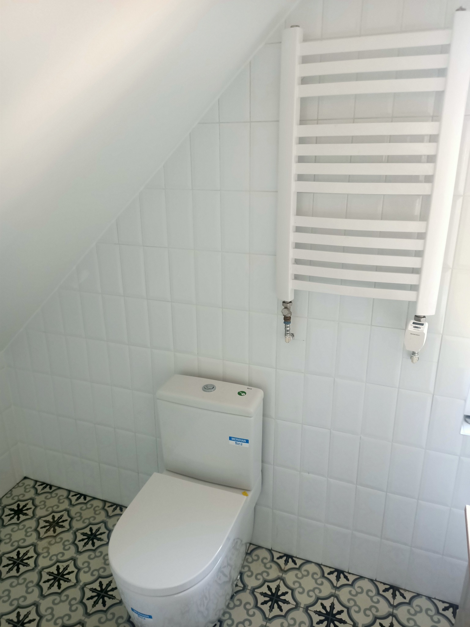 Biała toaleta kompaktowa z dekoracyjną podłogą w łazience na poddaszu, obok grzejnik drabinkowy.