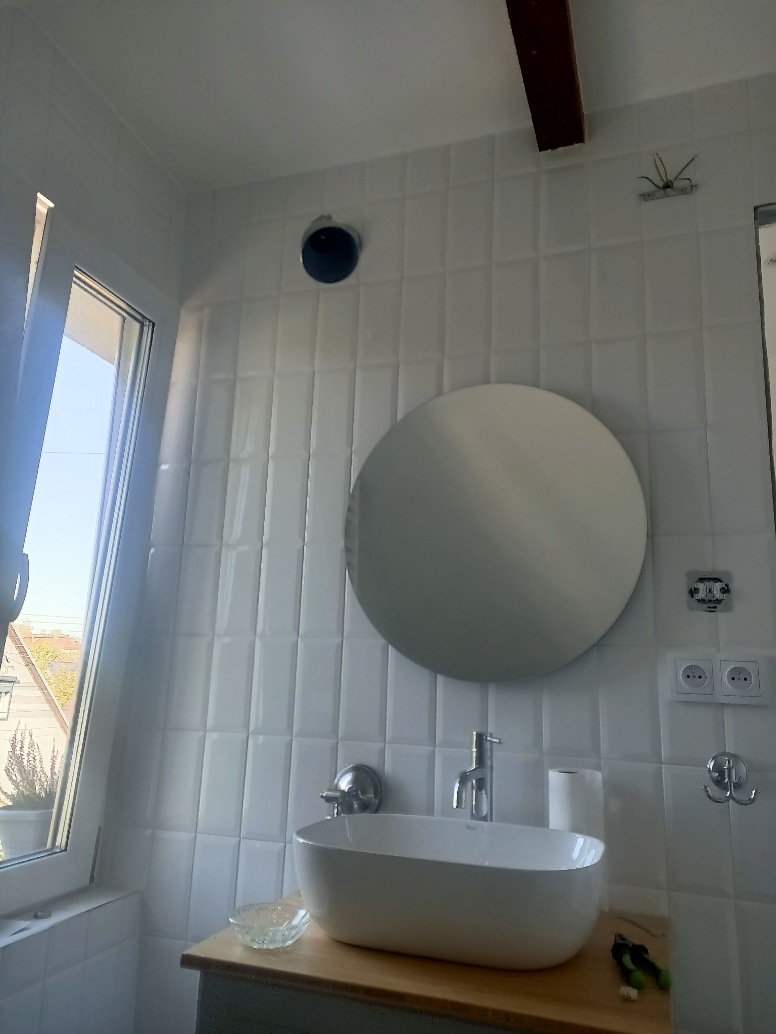 Nowoczesna łazienka z owalną umywalką nablatową, okrągłym lustrem i białymi, pionowo ułożonymi płytkami imitującymi cegłę, drewniany blat pod umywalką, widoczne gniazdka elektryczne i wylot...