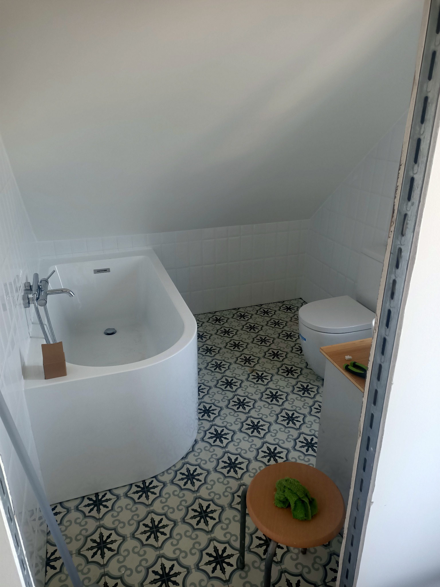 Biała wanna z chromowaną armaturą, biała toaleta i umywalka w małej łazience na poddaszu z wzorzystymi płytkami podłogowymi. Na taborecie leży zielona ściereczka.