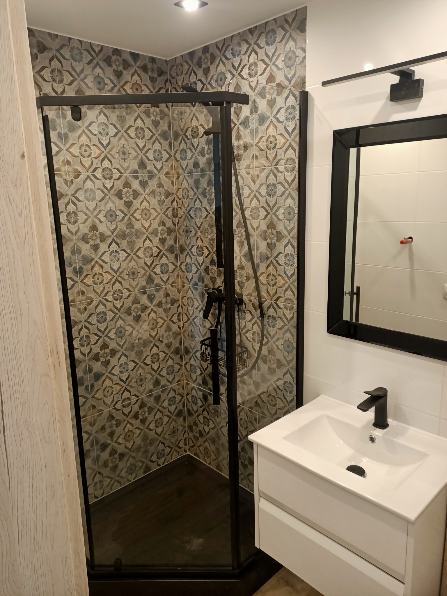 Wykończona łazienka z kabiną prysznicową z czarnymi ramami i wzorzystymi płytkami, biała umywalka z czarną baterią, czarne ramy lustra i oświetlenie.