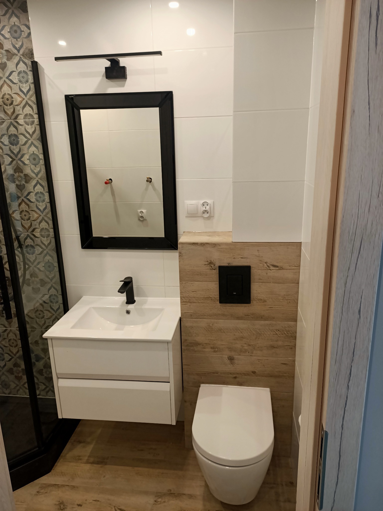 Nowoczesna łazienka z białymi płytkami, drewnopodobnymi panelami, wiszącą toaletą i czarną armaturą, widok na umywalkę z szafką i lustro z oświetleniem.