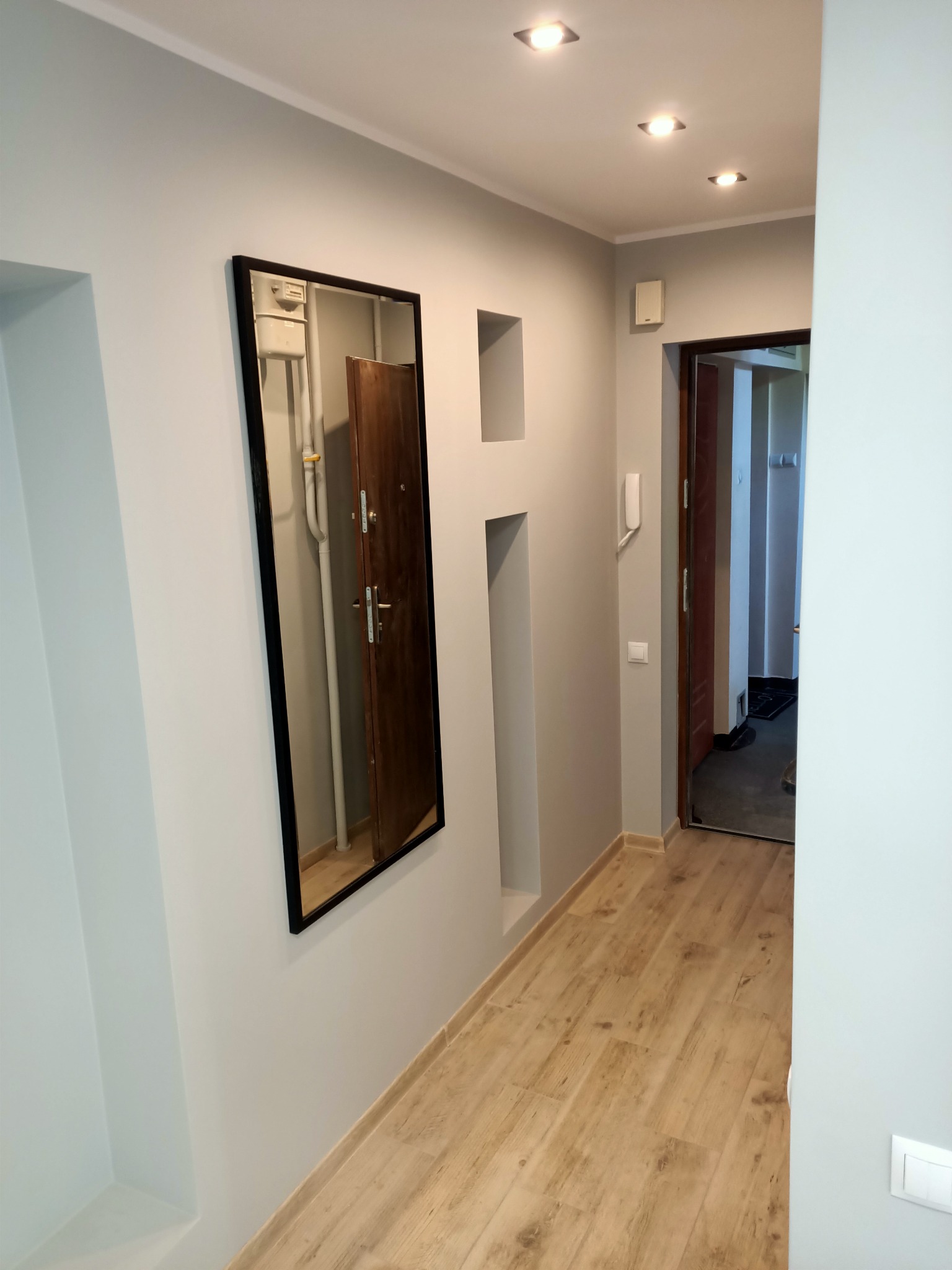 Szary korytarz z drewnianą podłogą, lustrem odbijającym drzwi wejściowe i wnękami w ścianie.