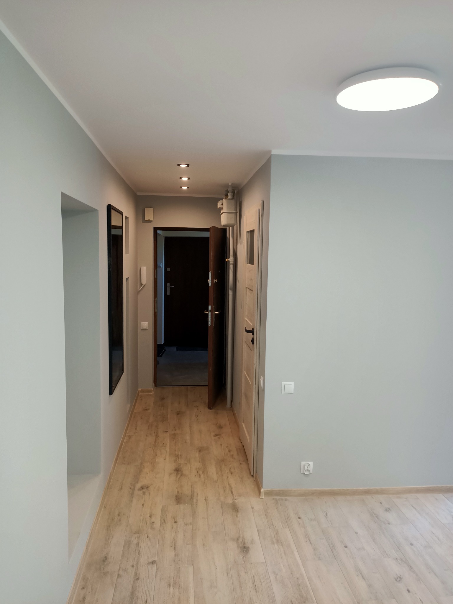 Wnętrze mieszkania po remoncie: jasnoszare ściany, panele podłogowe w odcieniach drewna, widok na korytarz z oświetleniem punktowym i otwartymi drzwiami.
