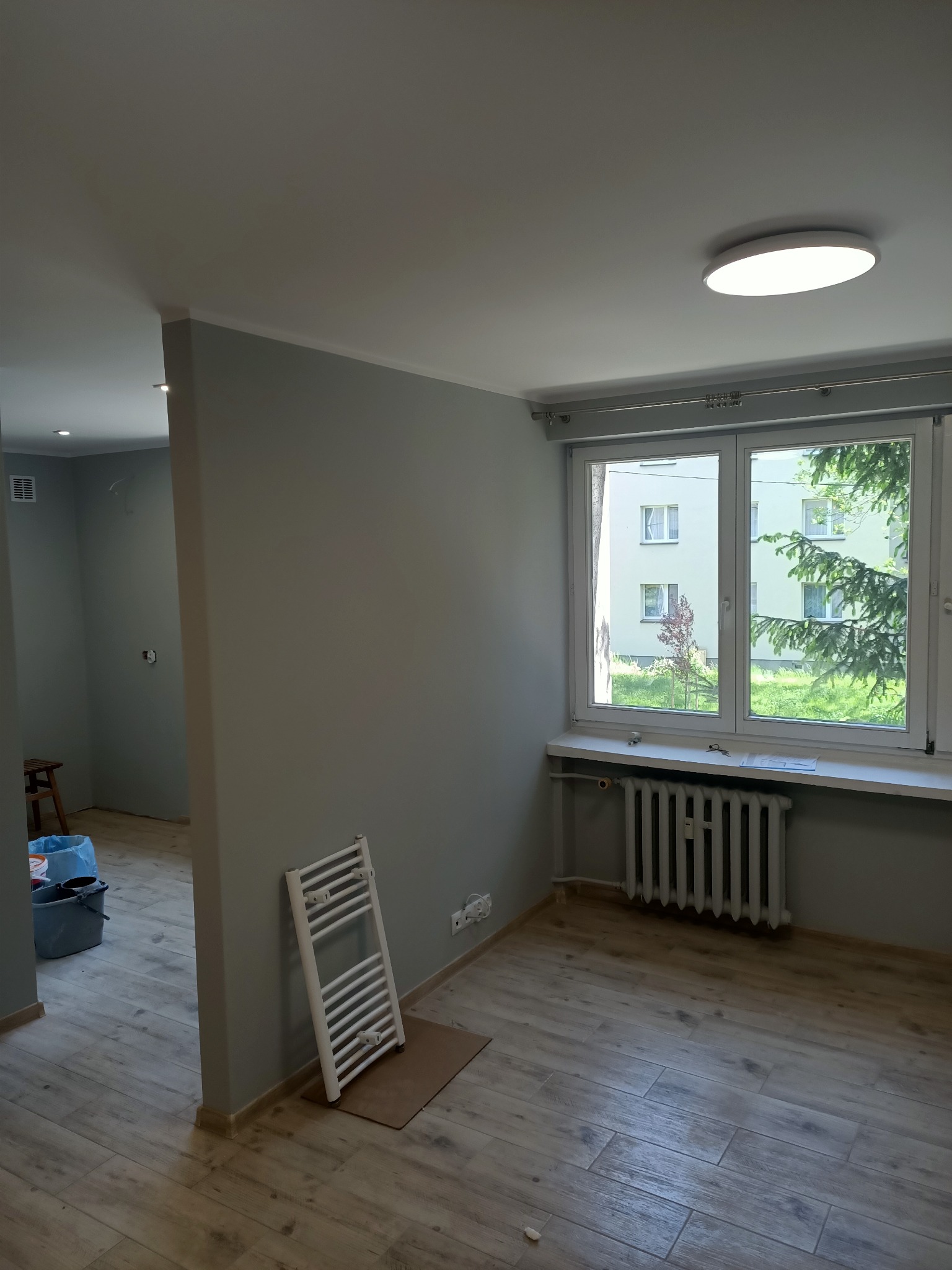 Wnętrze pokoju po odświeżeniu: szare ściany, drewnopodobne panele podłogowe, biały grzejnik, okno z widokiem na zieleń, minimalistyczna lampa sufitowa, drabinka grzejnikowa oparta o ścianę.