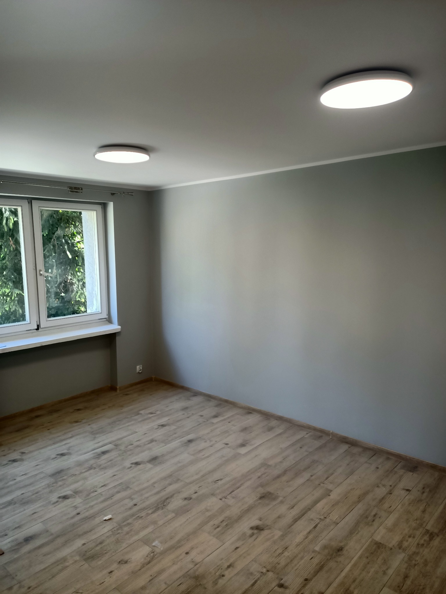 Pomalowane na szaro puste pomieszczenie z panelami podłogowymi, oświetlone dwoma okrągłymi lampami sufitowymi i naturalnym światłem wpadającym przez okno.