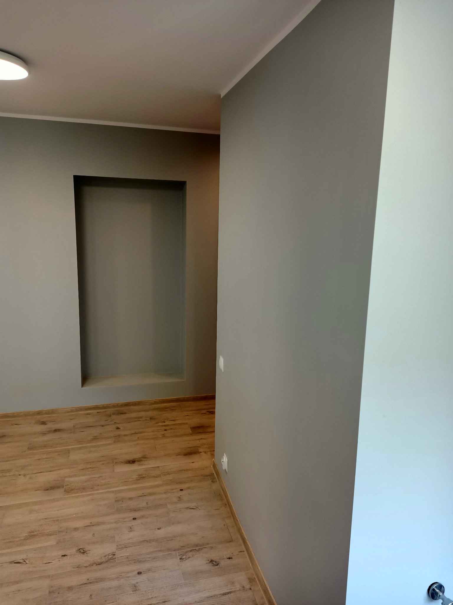 Wnętrze pokoju po remoncie: szare ściany, drewnopodobne panele podłogowe, wnęka w ścianie pomalowana na szaro, widoczne listwy przypodłogowe.