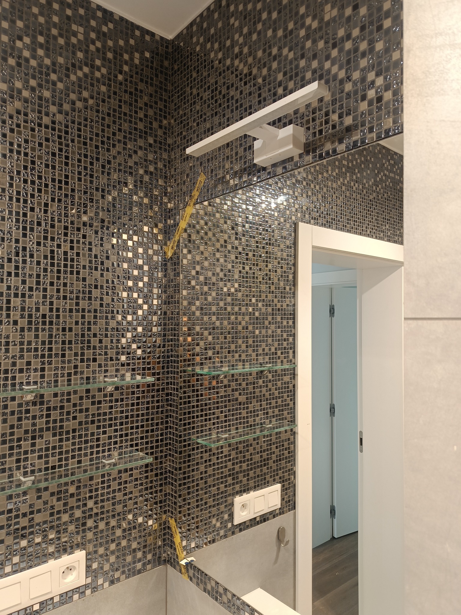Mozaika z drobnych, błyszczących płytek w odcieniach szarości i srebra, szklane półki, oświetlenie nad lustrem oraz widoczne gniazdka elektryczne w łazience, z fragmentem żółtej taśmy malarskiej.