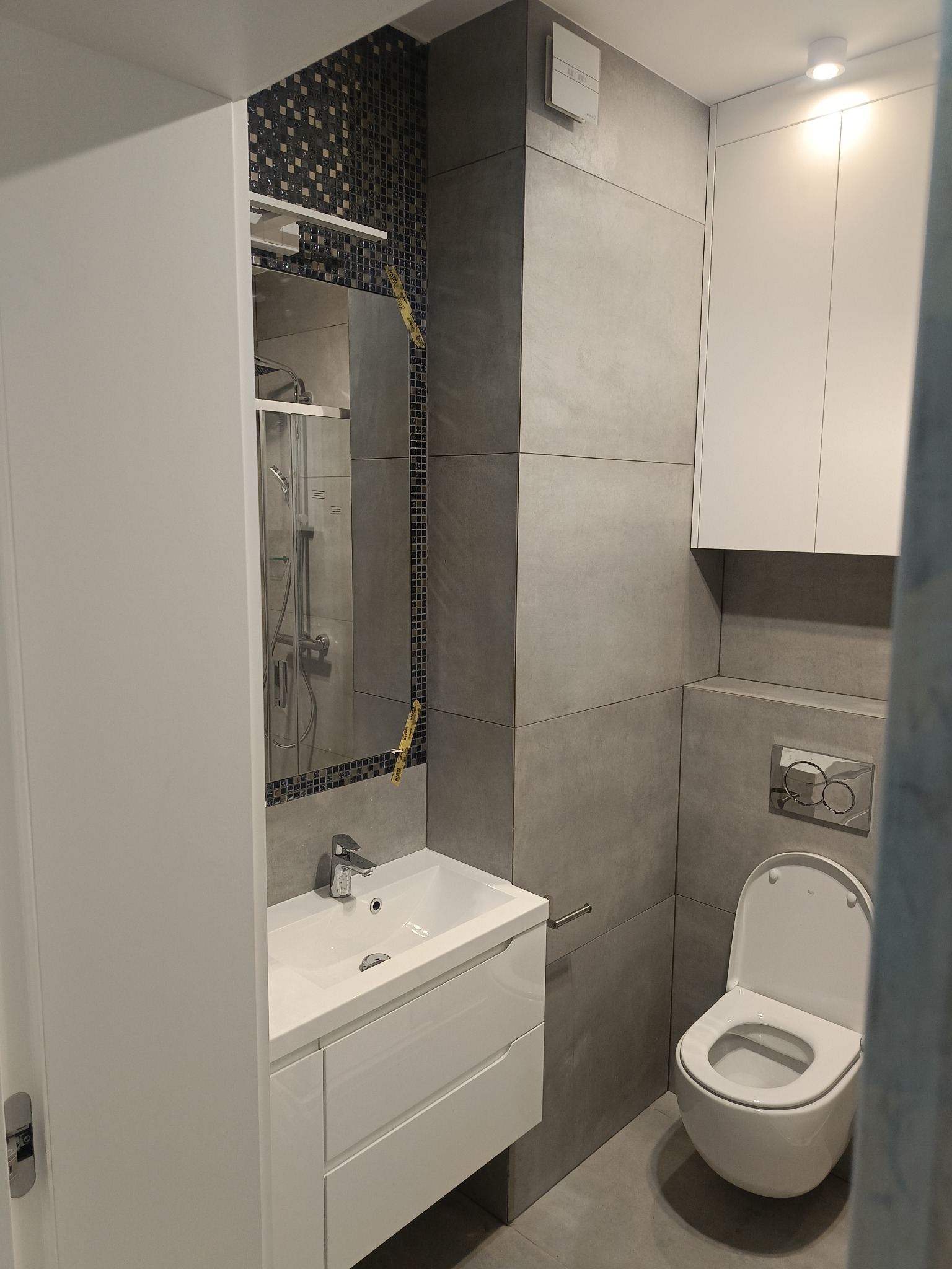 Nowoczesna łazienka z szarymi płytkami, białą umywalką z szafką, toaletą i lustrem z mozaiką, oświetlona punktowo.