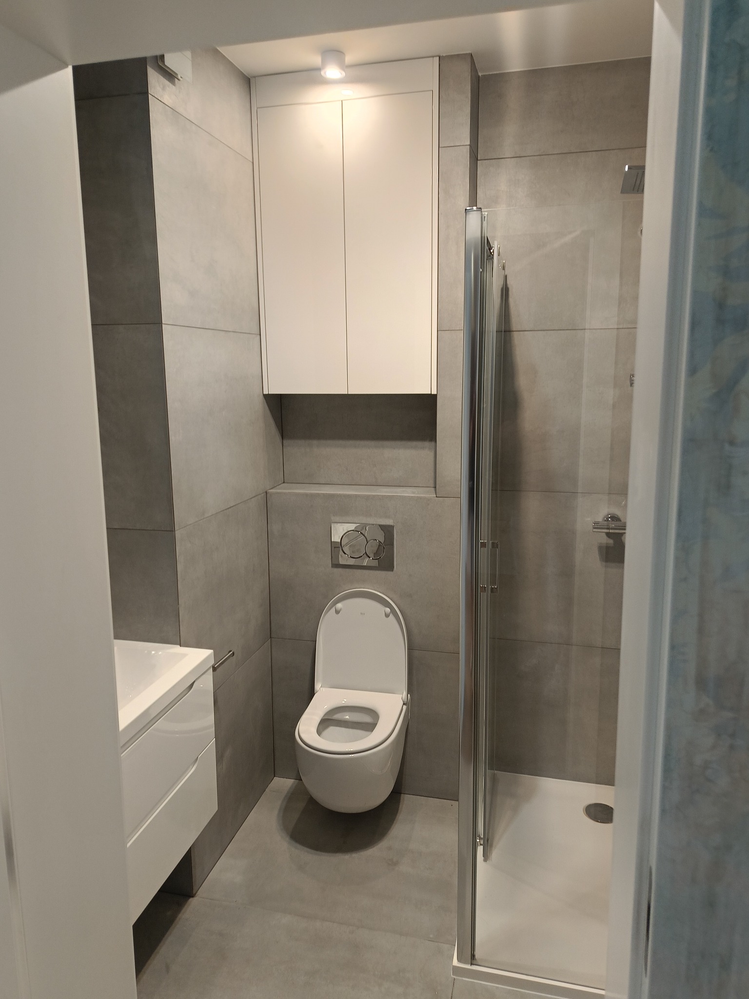 Nowoczesna łazienka w odcieniach szarości z białymi meblami: wisząca szafka, umywalka z szafką i toaleta, kabina prysznicowa z metalową ramą.