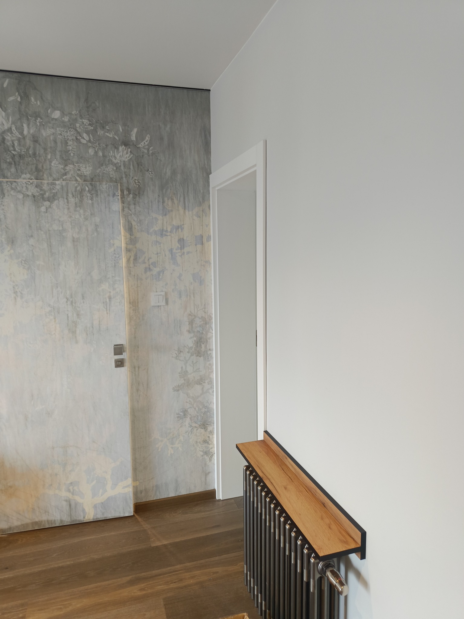 Wnętrze mieszkania z dekoracyjną ścianą z motywem roślinnym, grzejnikiem z drewnianą półką i parkietem