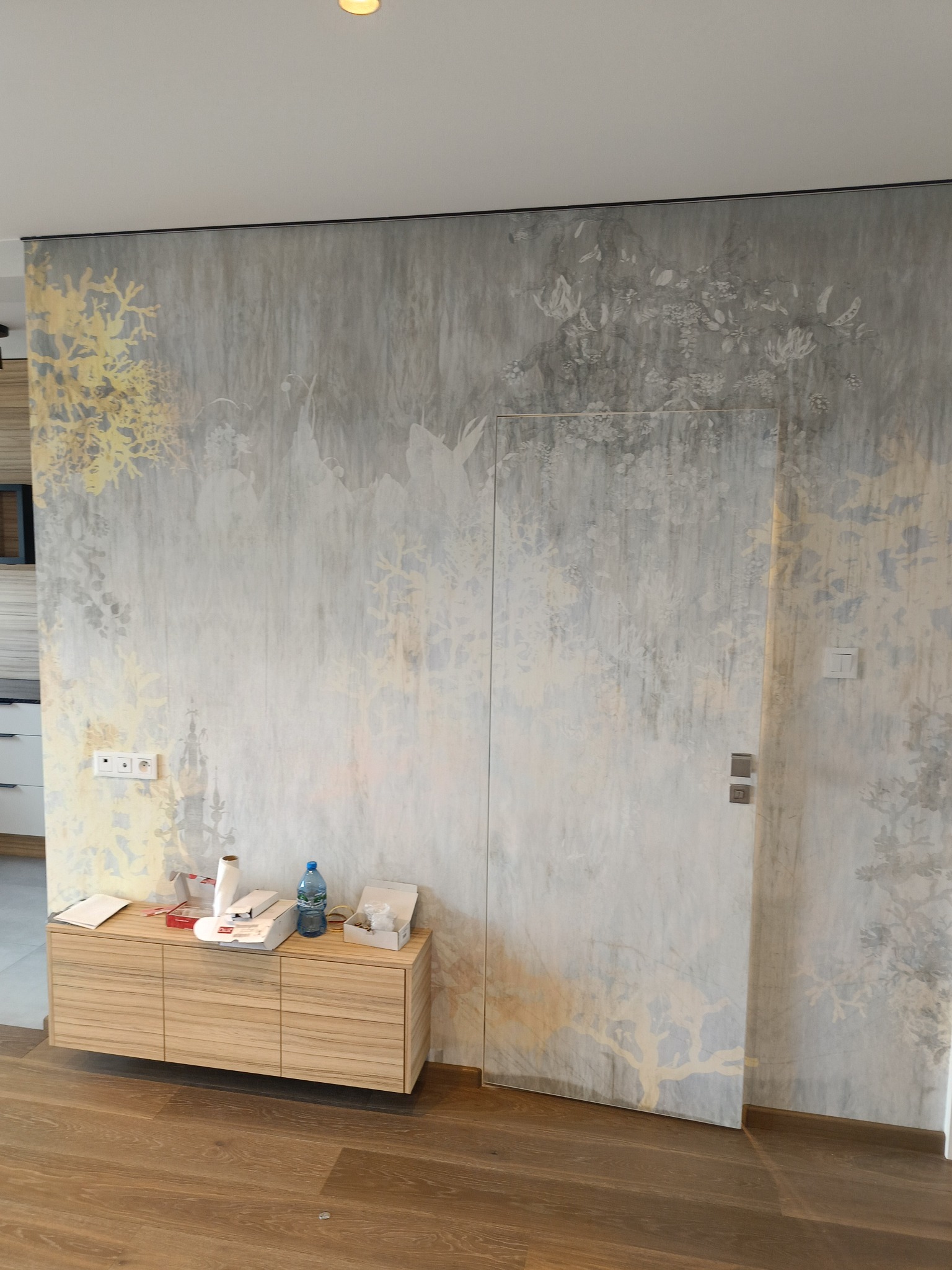 Ściana z ukrytymi drzwiami pokryta szarą tapetą z motywem roślinnym w odcieniach złota i bieli, z drewnianą szafką i akcesoriami malarskimi u dołu.