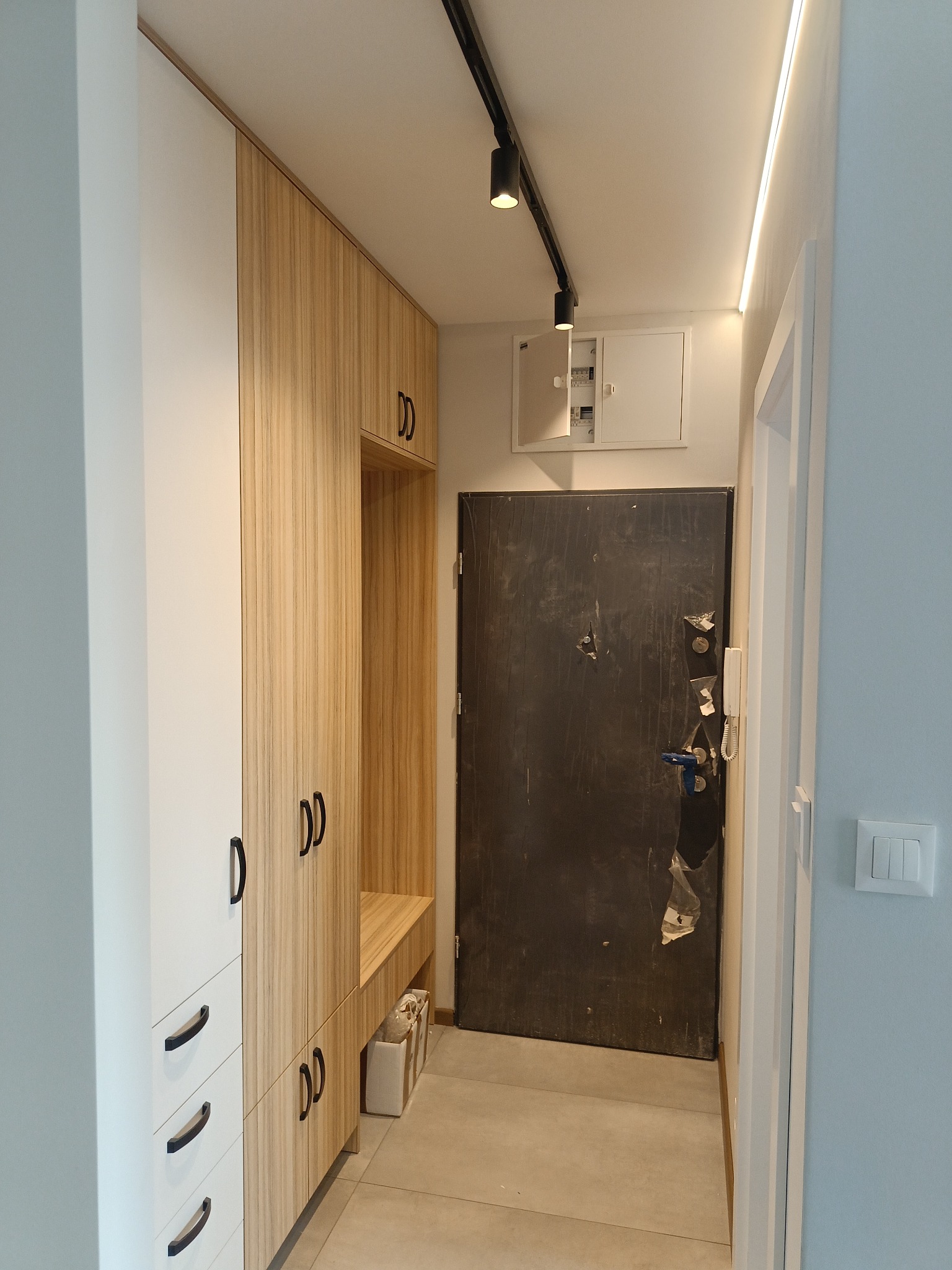 Przedpokój z zabudową szafy w odcieniach bieli i drewna, oświetlenie szynowe i listwa LED na suficie, drzwi wejściowe zabezpieczone folią.