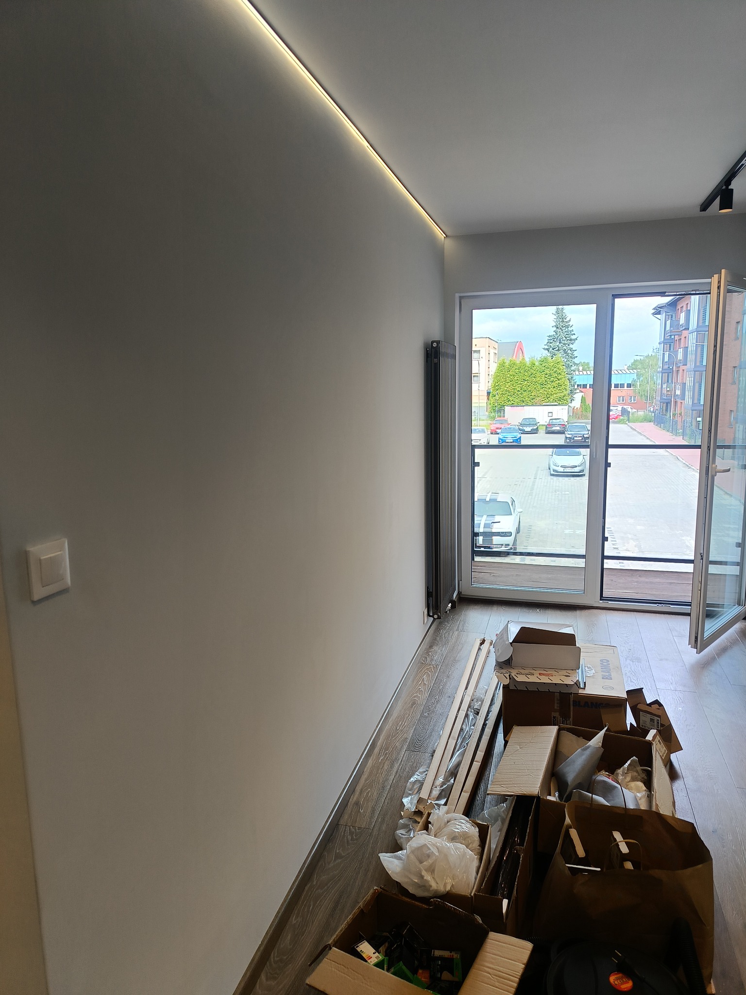 Wnętrze mieszkania w trakcie prac wykończeniowych: gładka, pomalowana ściana z oświetleniem LED podsufitowym, widok na zewnątrz przez balkonowe okno, na podłodze kartony i materiały budowlane.