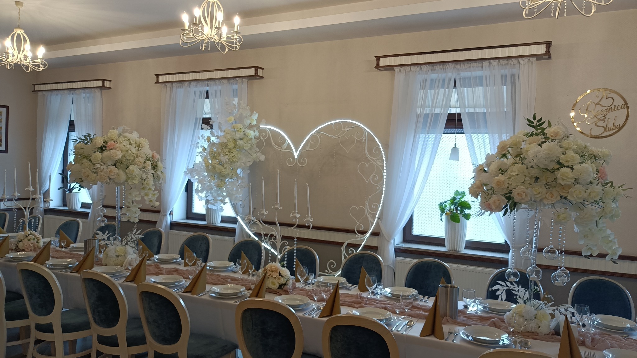 Uroczysty stół weselny z białym obrusem, dekoracjami kwiatowymi w wysokich wazonach, złotymi serwetkami i podświetlanym sercem z ornamentami.
